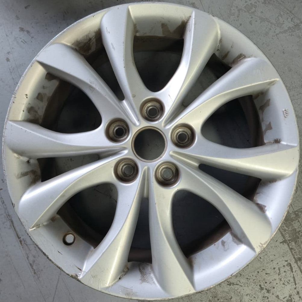 A used silver alloy car wheel rim with a multi-spoke design, showing some dirt and wear, photographed on a concrete floor.