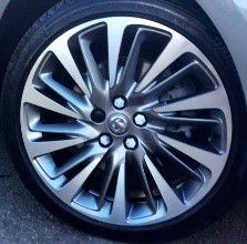 A close-up of a multi-spoke alloy wheel with a silver and dark gray finish, fitted with a black tire. The center cap features a circular emblem.