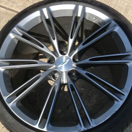 A close-up of a silver multi-spoke alloy car wheel with a black tire, photographed outdoors on a concrete surface. The center cap features the Volvo logo.