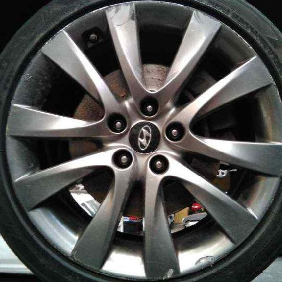 A close-up of a silver multi-spoke alloy car wheel with a black tire, showing the center cap logo and brake disc behind it.