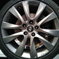 A close-up of a silver multi-spoke alloy car wheel with a black tire, showing the center cap logo and brake disc behind it.