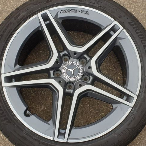 A close-up of a Mercedes-AMG wheel rim with a dark gray and silver multi-spoke design, fitted with a black tire. The Mercedes star logo is visible in the center, and 'AMG' is written on the outer lip.