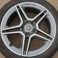 A close-up of a Mercedes-AMG wheel rim with a dark gray and silver multi-spoke design, fitted with a black tire. The Mercedes star logo is visible in the center, and 'AMG' is written on the outer lip.